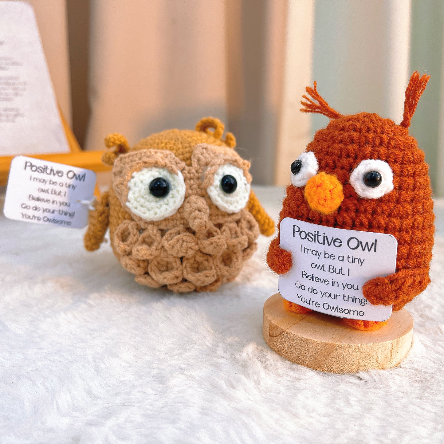 Two crochet owls, one brown and one orange, on a white surface with a blurred background.