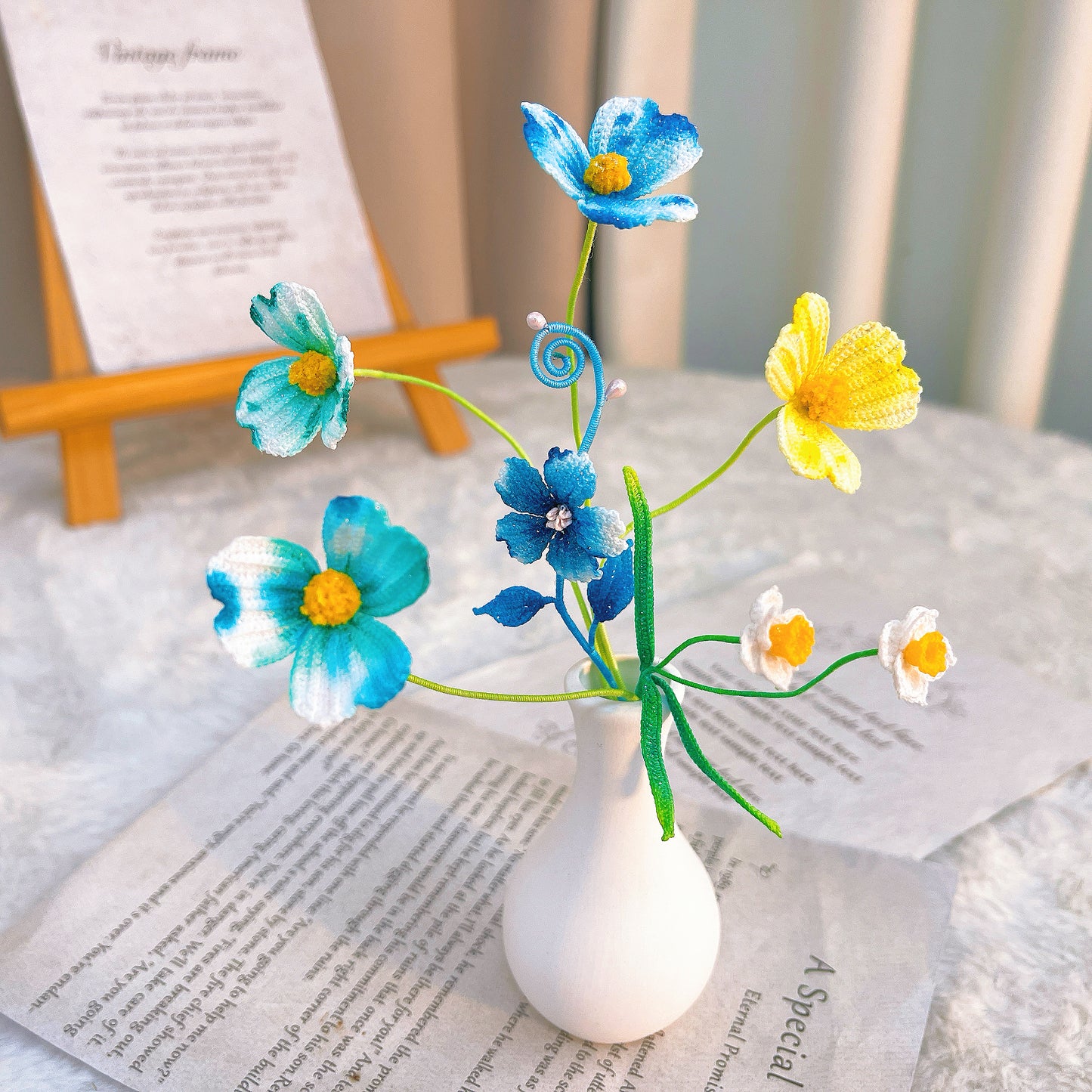 Small white vase with blue, yellow, and white flowers on a surface with text.