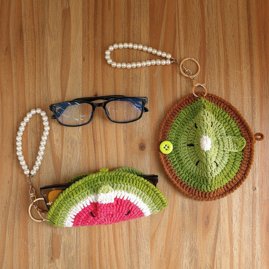 Two crocheted sunglasses pouches styled on a white faux fur background, one shaped like a watermelon slice and one like a kiwi fruit, both with pearl bead wristlets and gold-tone keychains. A pair of black-framed glasses is also displayed.