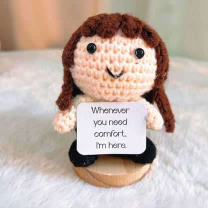 A crocheted doll with brown hair, a cream face, and black clothing sits on a small wooden base, holding a white sign that reads, 'Whenever you need comfort, I'm here.'.