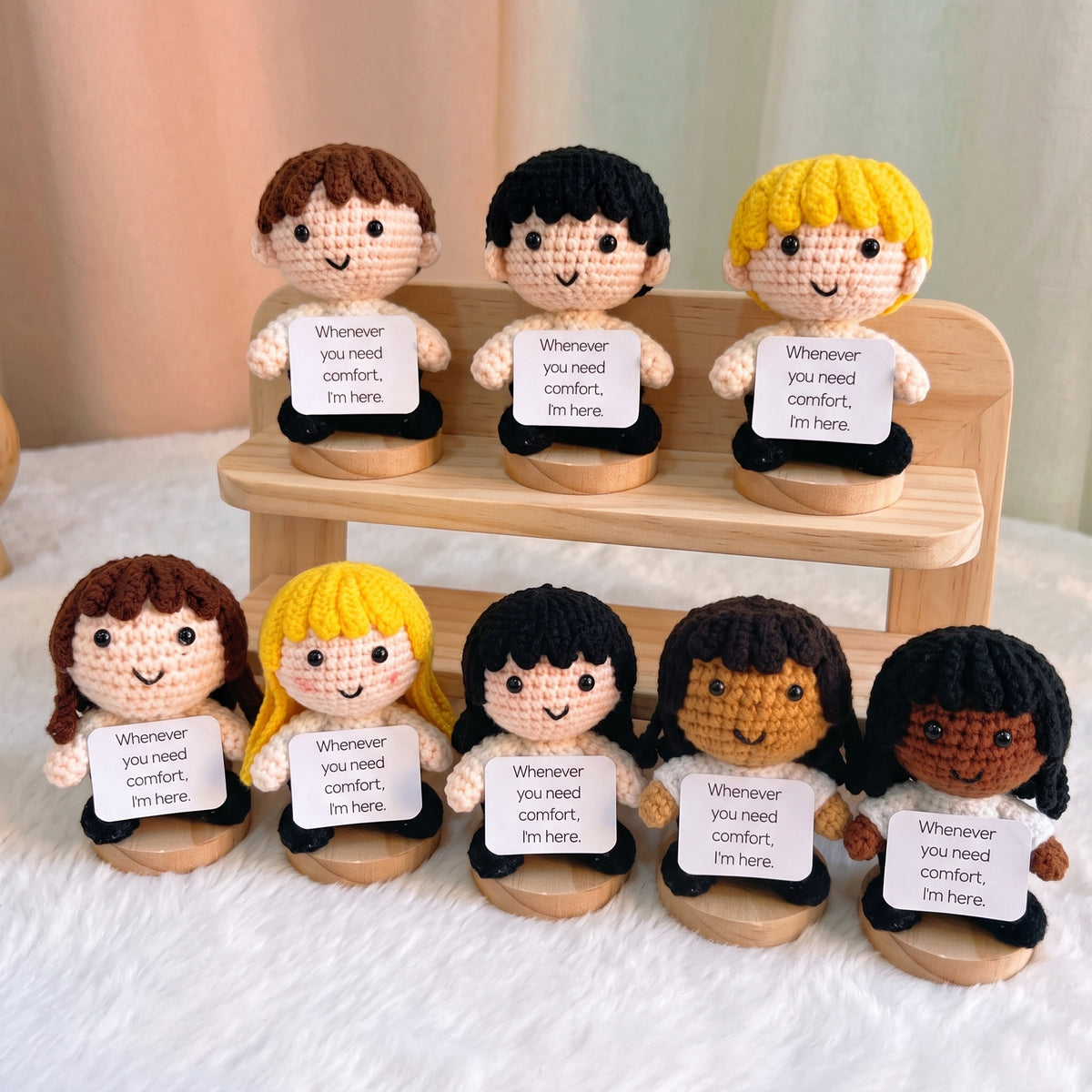 Seven crocheted comfort dolls with different hair colors and skin tones, each holding a white sign that reads 'Whenever you need comfort, I'm here.' They are arranged on a small wooden display stand and a white textured surface against a soft beige background.