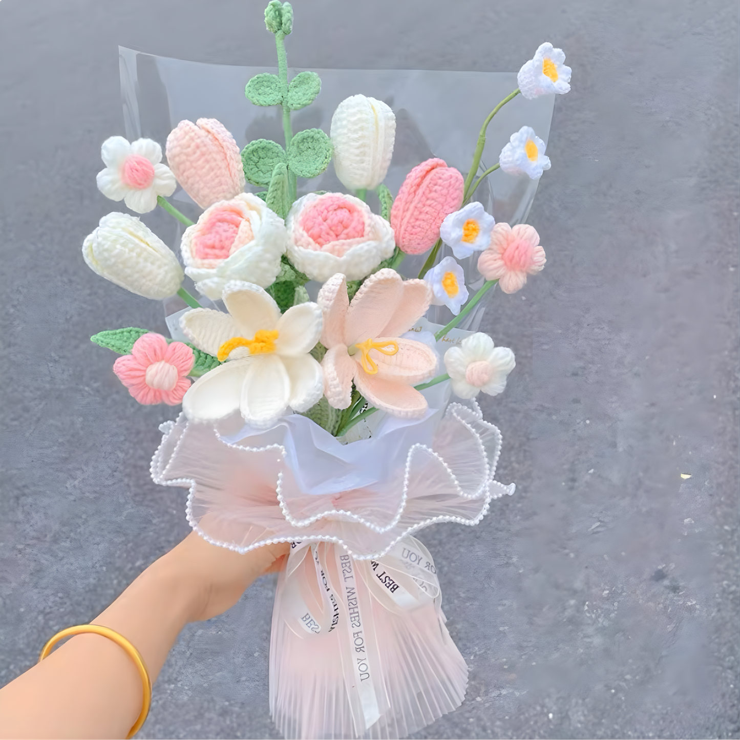 Bouquet of knitted flowers wrapped in sheer paper with a bow, held against a gray background.