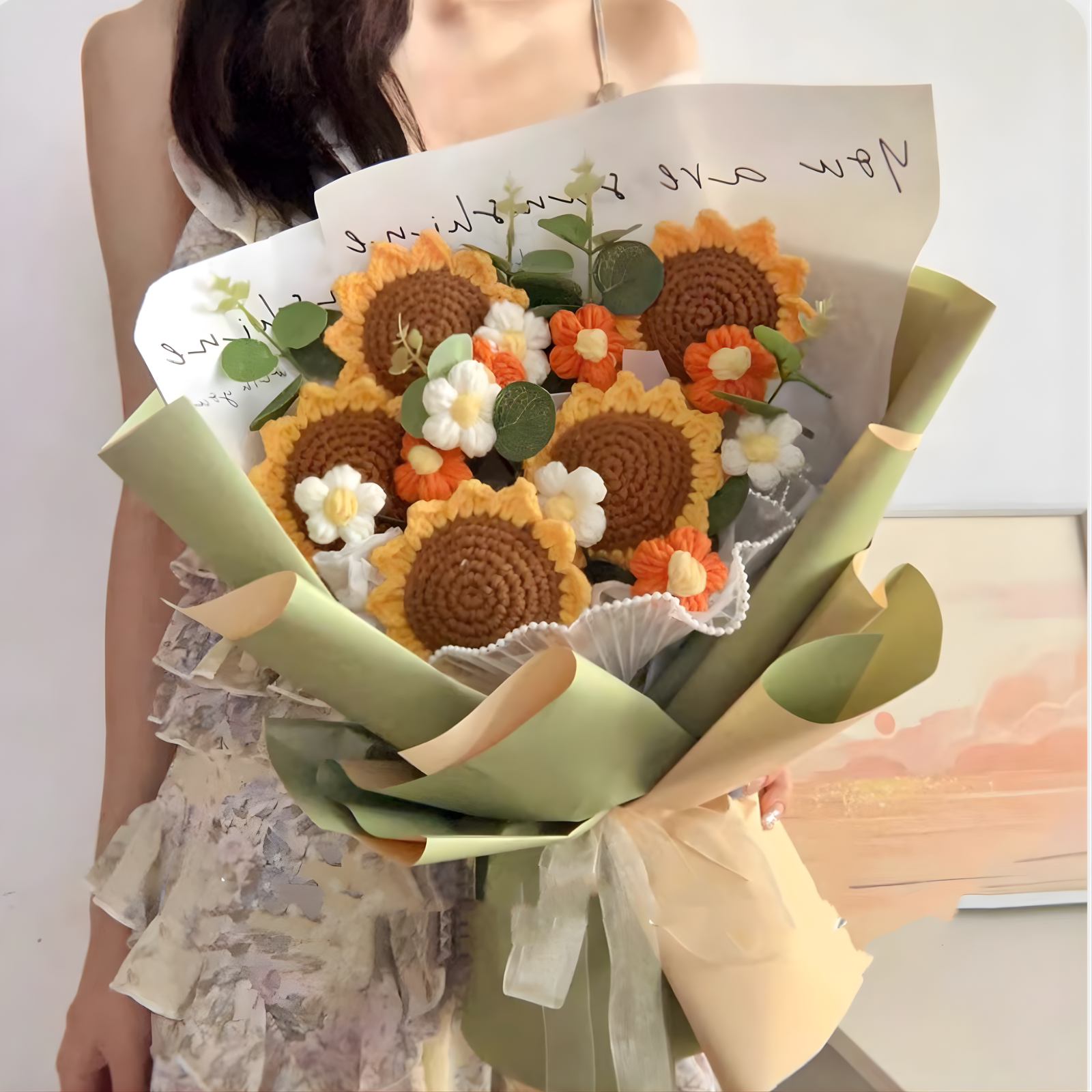 Bouquet of knitted sunflowers and white flowers with a card held by a person.