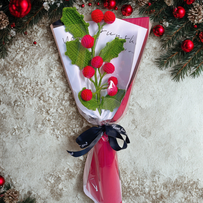 Knitted holly branch with red berries on a wrapped gift against a textured surface with Christmas decorations.