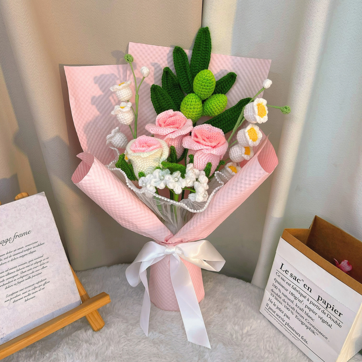 Handmade crochet flower bouquet in a pink wrapped vase with a white ribbon, styled on a white faux fur surface with books in the background.