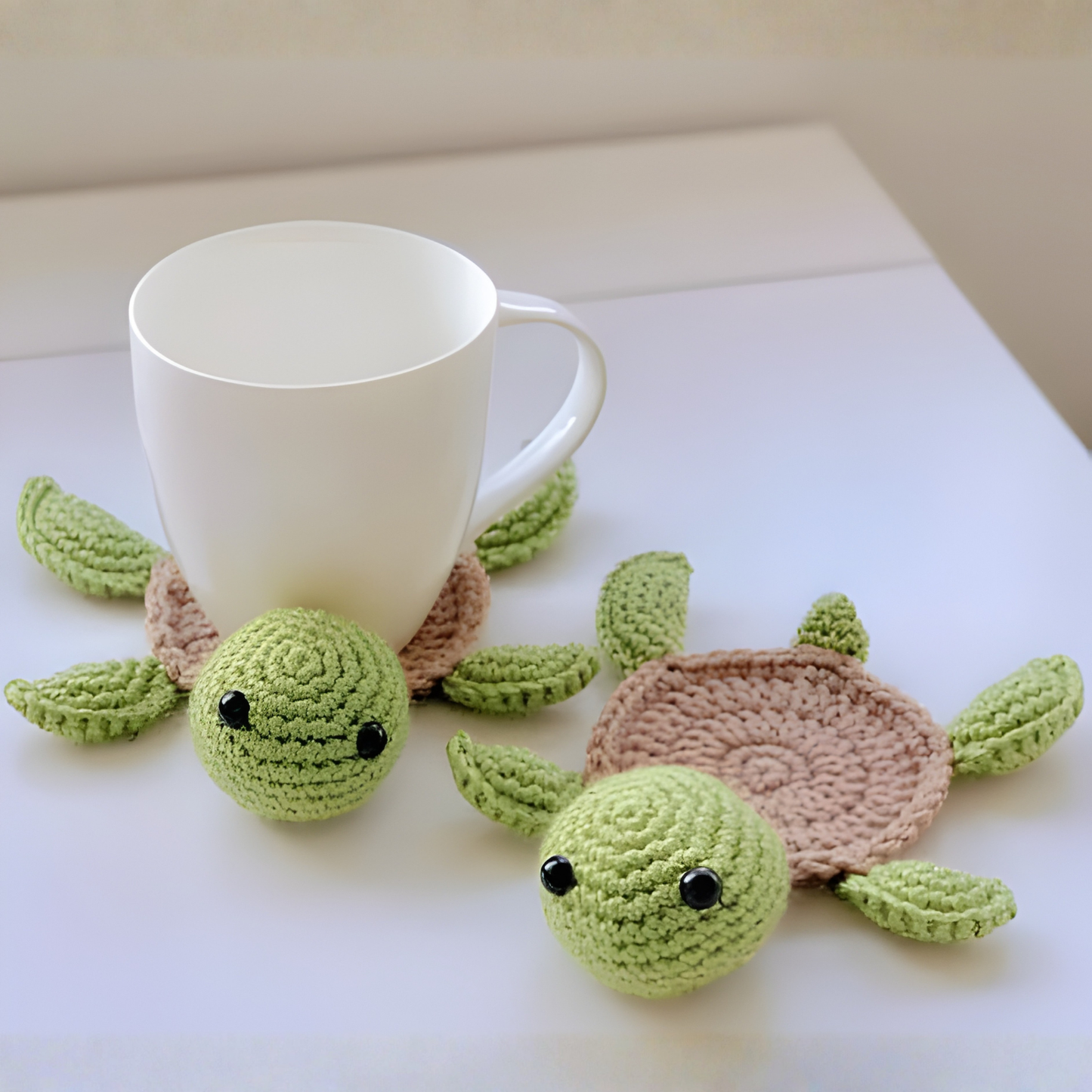 Two green turtle-shaped crochet coasters with a white mug on a light surface.